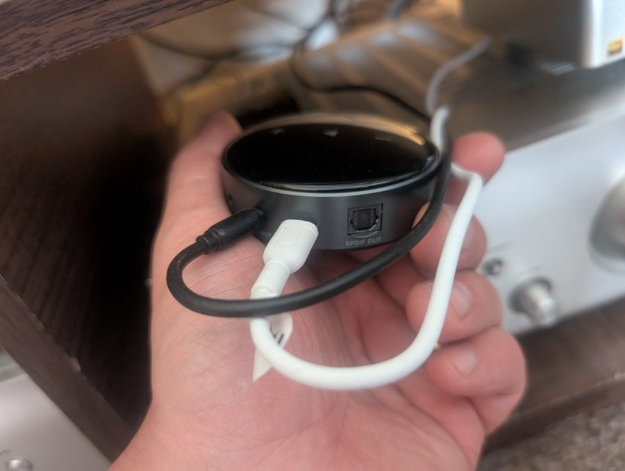 WiiM Mini streamer shown in hand with connected power and optical cables, highlighting its compact size and rear ports.